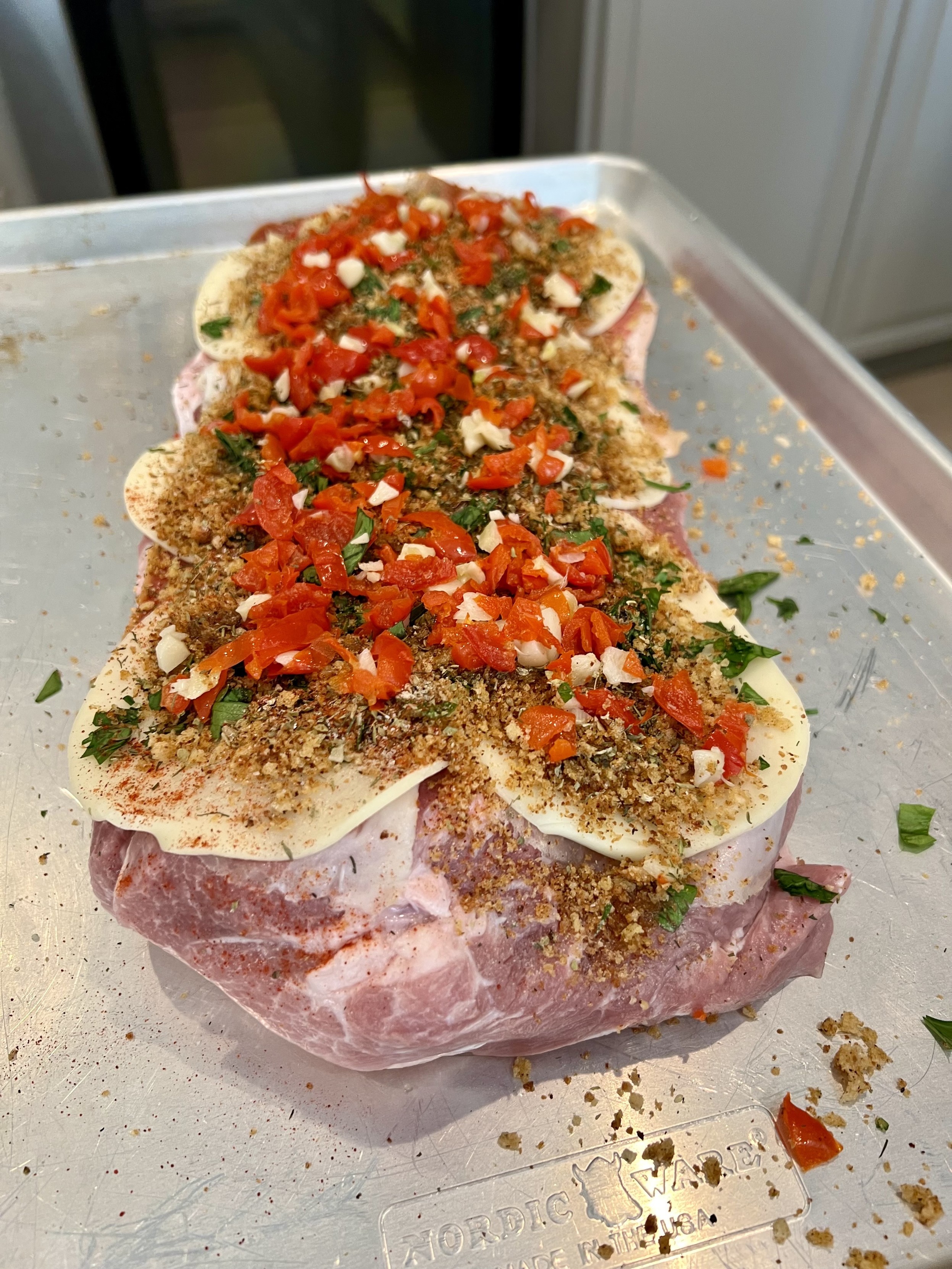 A seasoned piece of meat is displayed on a metal tray, topped with herbs, spices, chopped red peppers, and thin slices of cheese. The preparation hints at a flavorful dish ready for cooking.