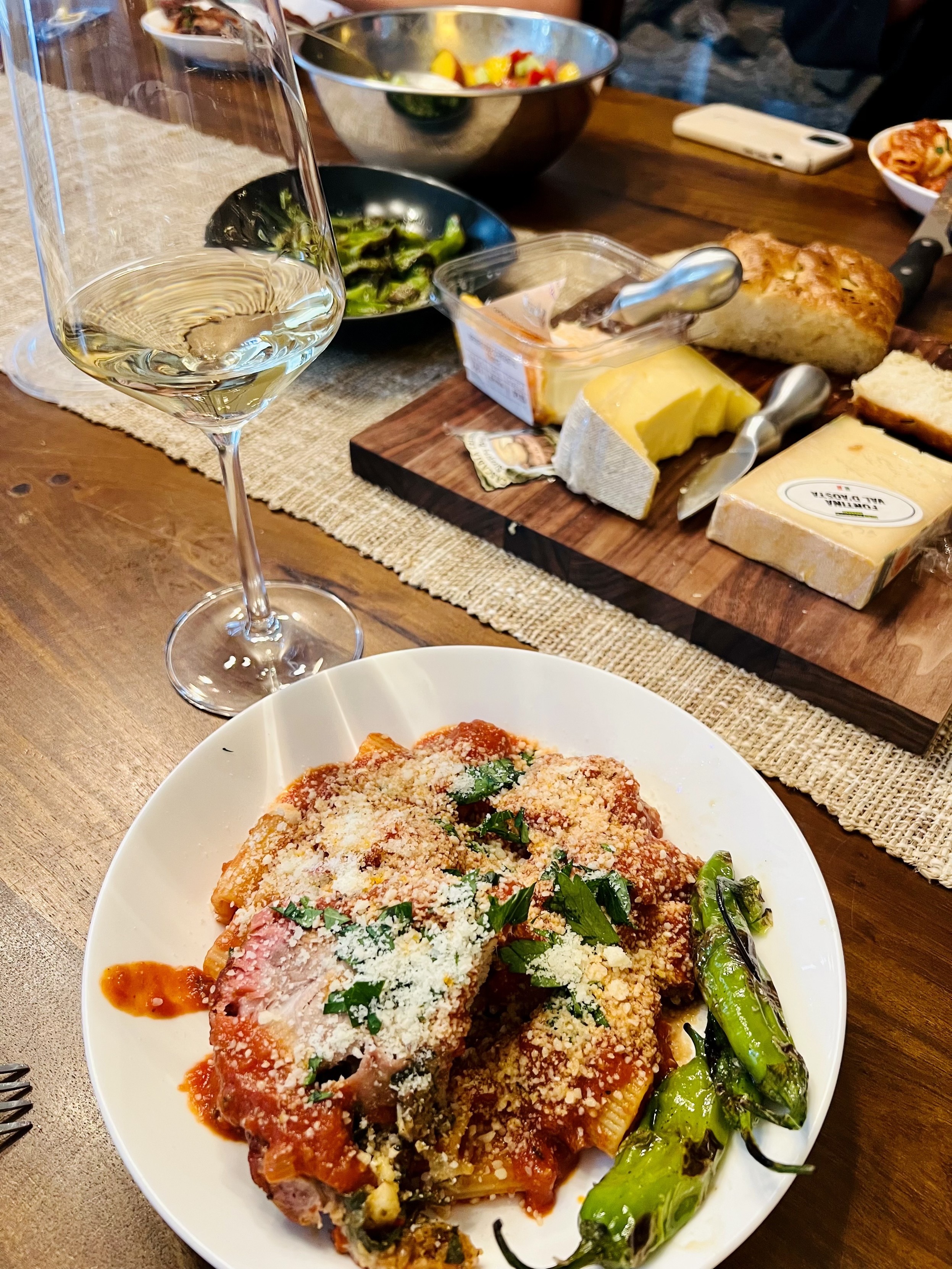 A plate of baked pasta with marinara sauce, topped with cheese and sprinkled herbs, accompanied by sautéed green peppers. A glass of white wine is beside it, with a wooden cheese board featuring various cheeses and bread in the background.