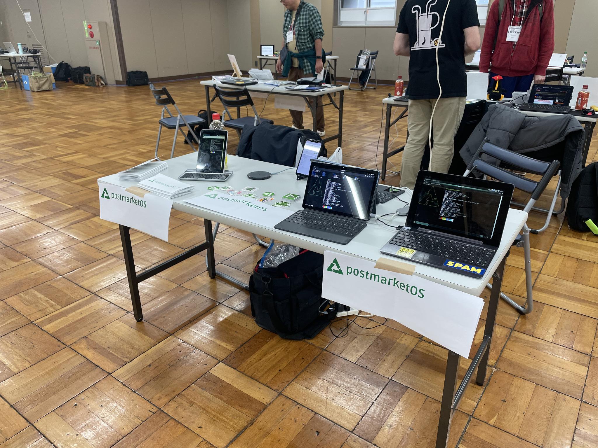 Wider shot showing the pmOS booth/table with 2 convertable devices, 1 phone, 1 tablet. leaflets and sticker ready for pick-up