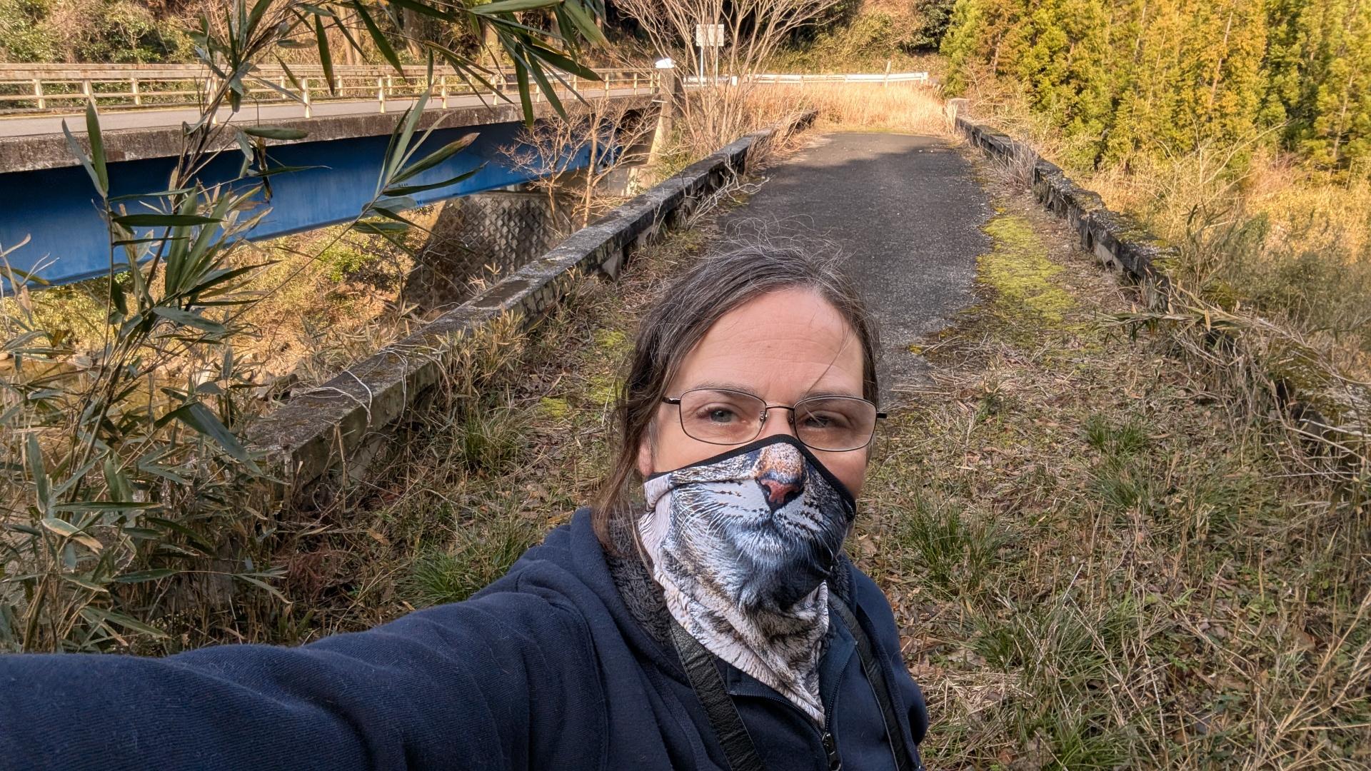 A man in a cat mask taking a selfie in front of an abandoned bridge. The new bridge is visable in the background.